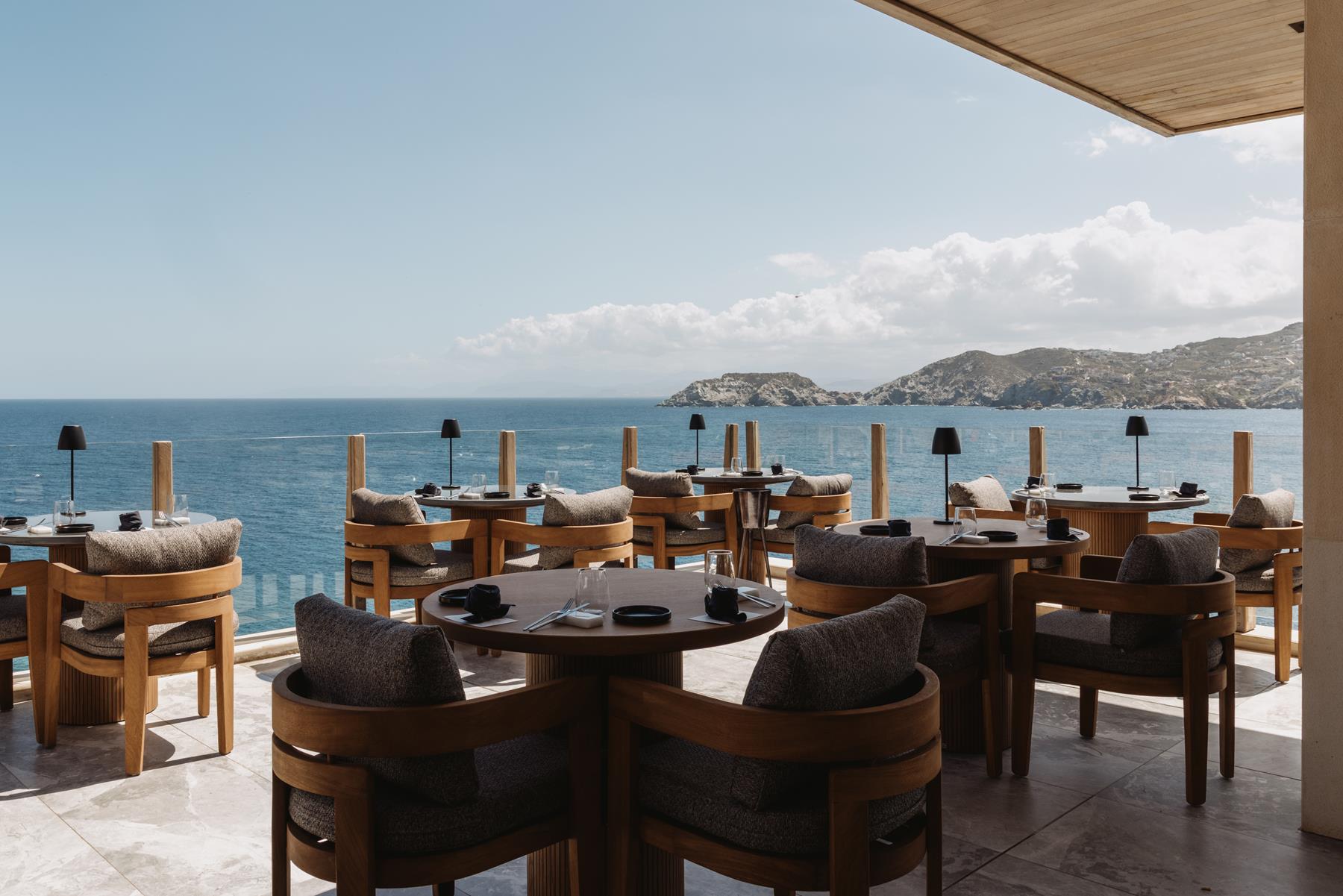 Oceano restaurant at Sea Side, round timber tables with dark lamps on a marble terrace facing the sea and distant Cretan headlands