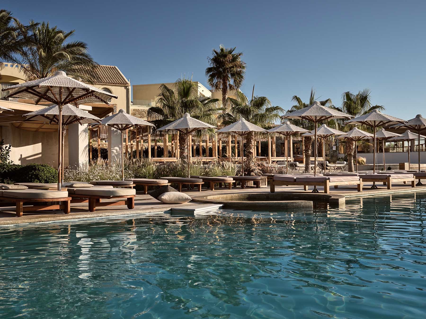 The poolside lounge at Sea Side, woven umbrellas, sun loungers and palm trees reflected in the calm pool water