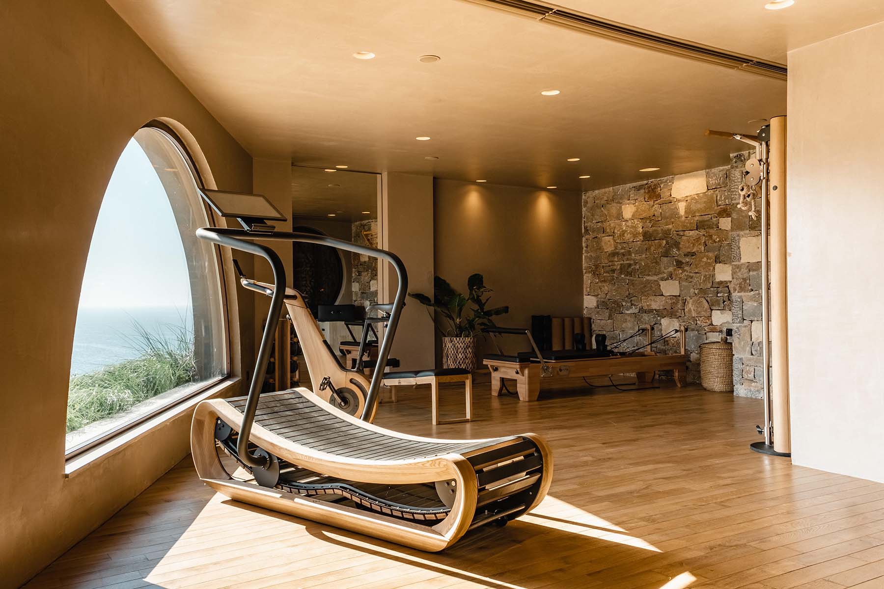 A curved wooden treadmill by the arched window of the Fitness House, gym equipment and sea view beyond